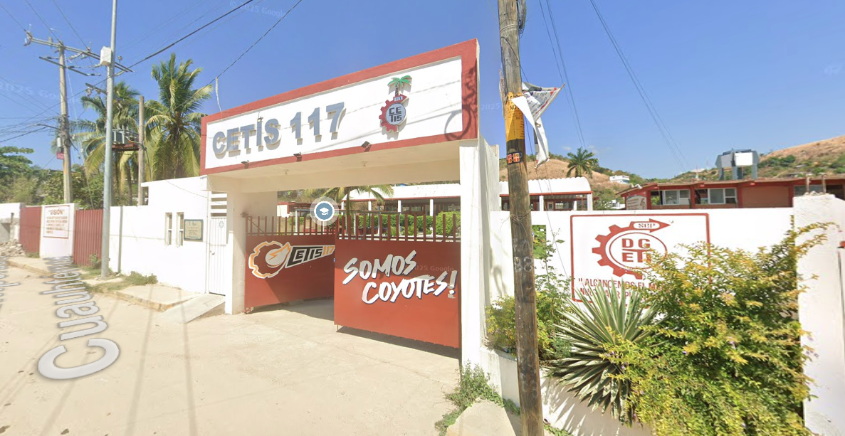 Fotografía en la que se muestra la entrada del plantel, conformada por un portón de color rojo en el que está escrito SOMOS COYOTES. Arriba del portón hay un muro blanco que en letras grandes dice CETIS 117. Las instalaciones están rodeadas por muros de color blanco.